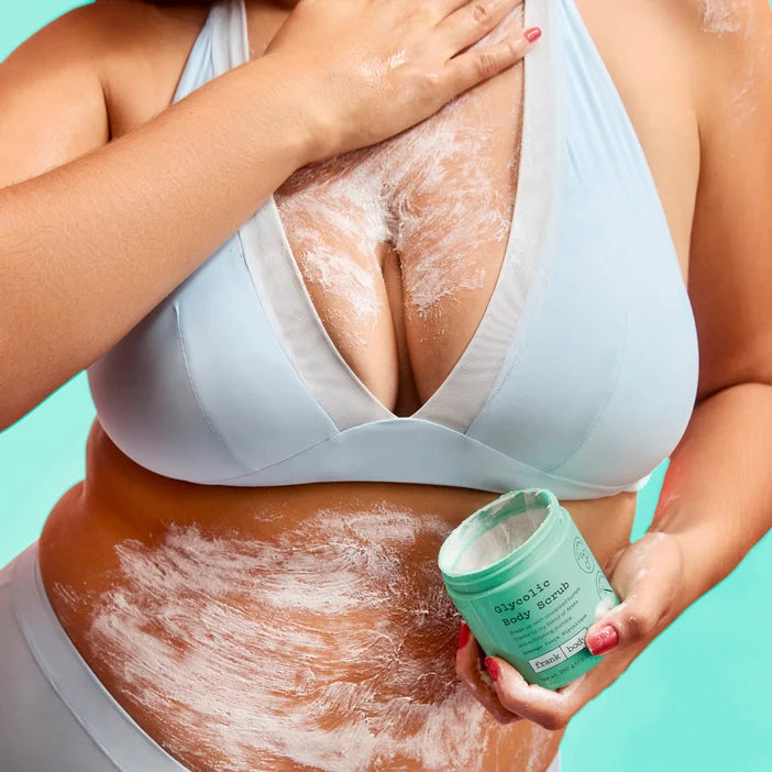 Person applying body scrub to their skin with a container of scrub in hand on a teal background