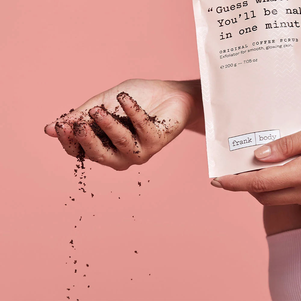 Hand holding coffee scrub with packaging labeled 'frank body' on a pink background