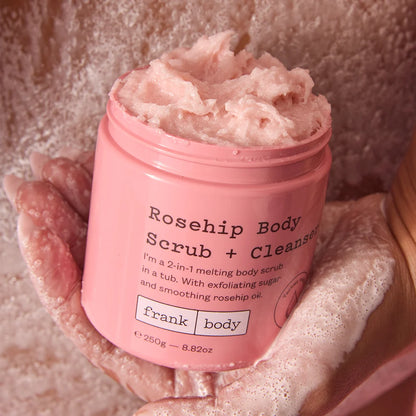 Pink jar of Rosehip Body Scrub + Cleanser by frank body with a hand holding it.