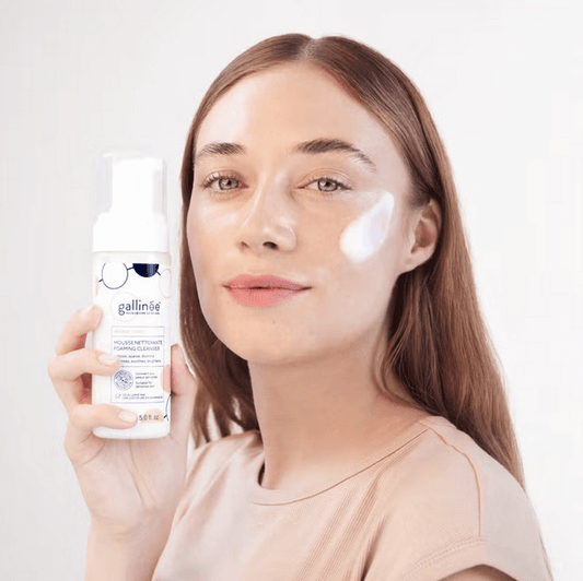 Woman holding Gallinée Prebiotic Foaming Facial Cleanser with foam on her cheek, showcasing skincare routine.