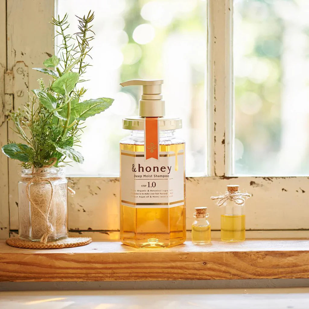 Bottle of '&honey' shampoo on a windowsill with plants and small bottles.
