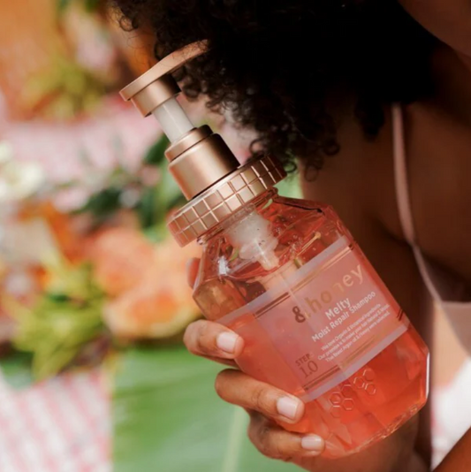 Hand holding a bottle of '&honey' hair product with a blurred floral background