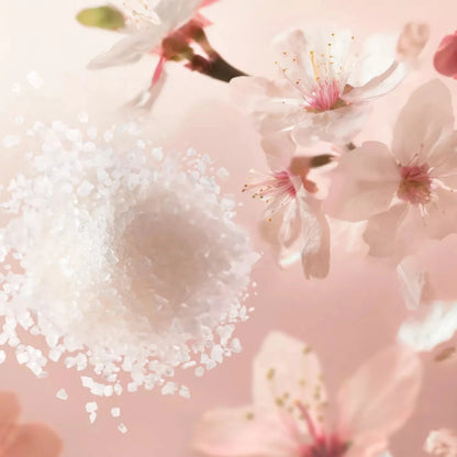 Close-up of a spoon with white powder surrounded by cherry blossoms on a pink background representing Rituals of Sakura Body Scrub