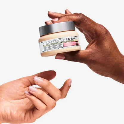 Hand holding a jar of IT Cosmetics  'Confidence in a Cream' product with another hand reaching out.