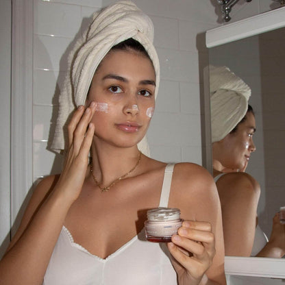 Woman applying Korres cream to her face with a towel on her head in front of a mirror.
