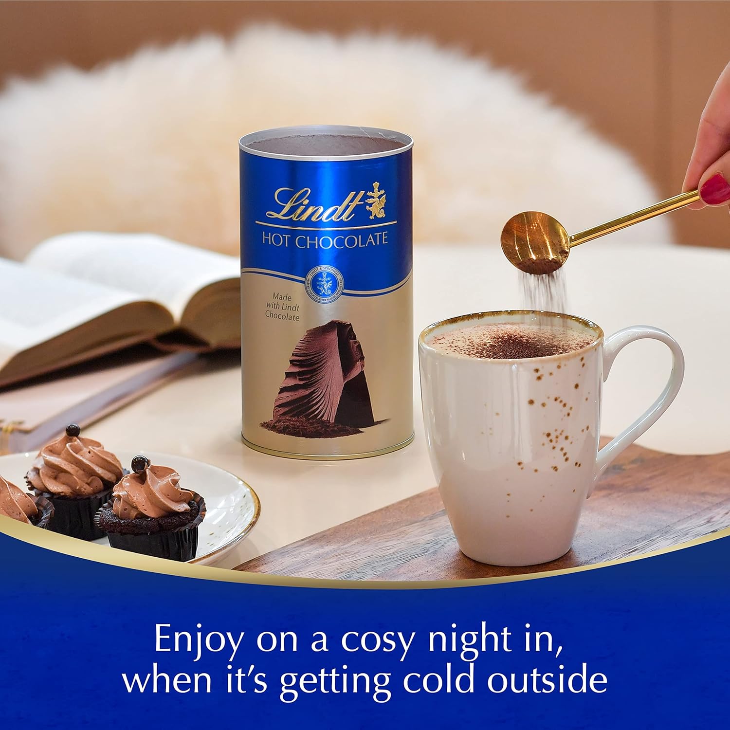 Lindt hot chocolate can with a mug being filled, surrounded by chocolate cupcakes and books on a cozy setting.