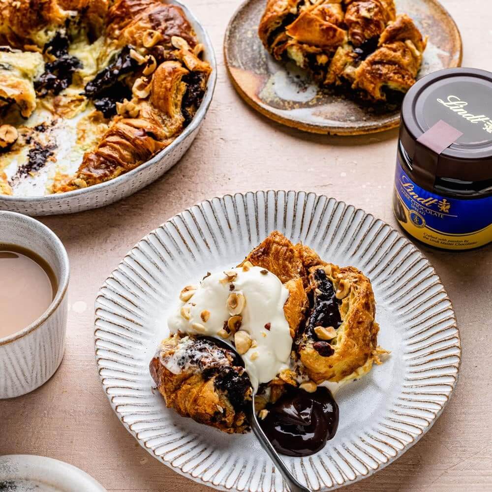 Sliced croissant with Lindt chocolate filling on a plate with a spoon, accompanied by a jar of jam and a cup of coffee.