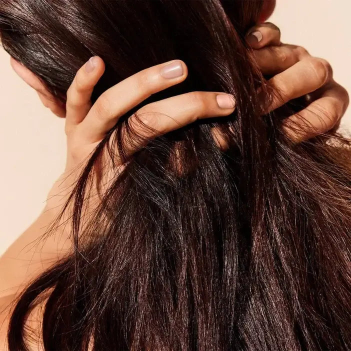 Close-up of a person's hand holding dark brown hair against a beige background Nuxe