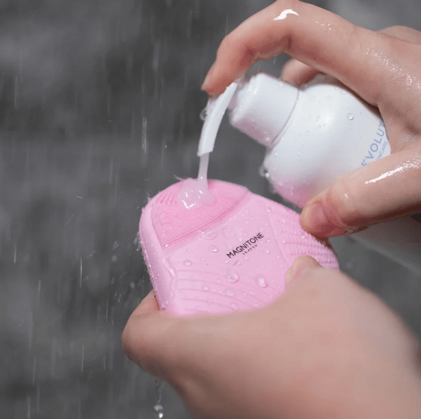 Hands holding Magnitone XOXO SoftTouch Silicone Cleansing Brush Pink with cleanser under running water.
