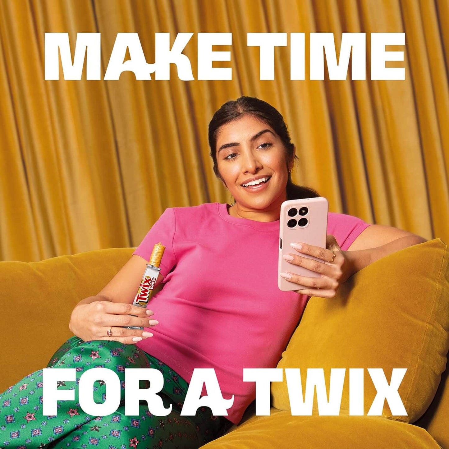 Woman sitting on a yellow couch holding a Twix bar and phone with text 'Make Time For A Twix' against a yellow curtain background.