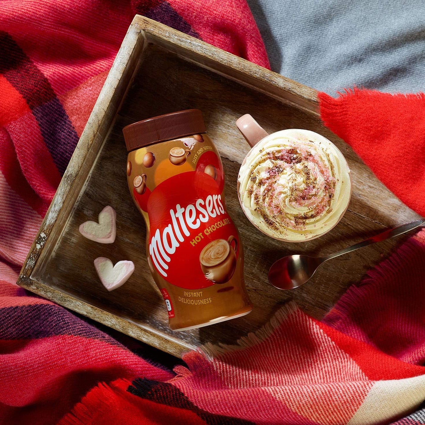 Maltesers hot chocolate mix and a cup of hot chocolate on a wooden tray with a red blanket underneath.