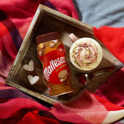 Maltesers hot chocolate mix and a cup of hot chocolate on a wooden tray with a red blanket underneath.