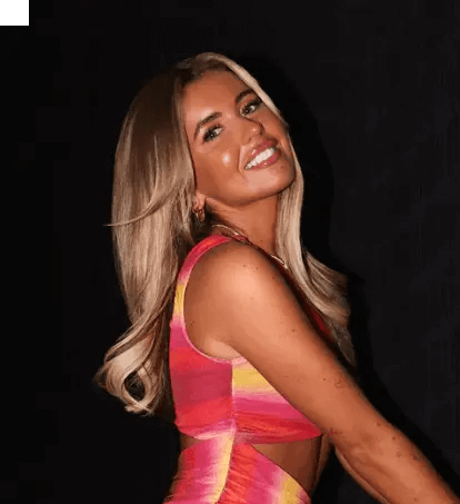 Smiling woman with long blonde hair wearing a colorful dress, posing against a dark background.