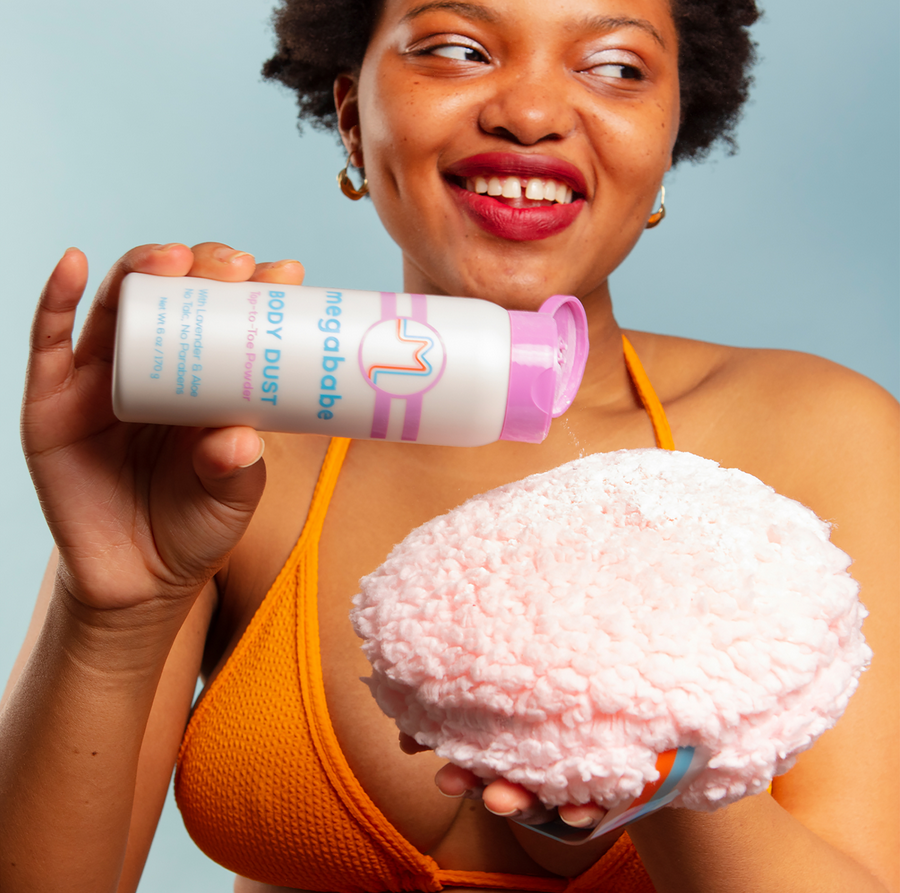 Woman holding a container of Megababe body dust and a fluffy applicator against a light blue background