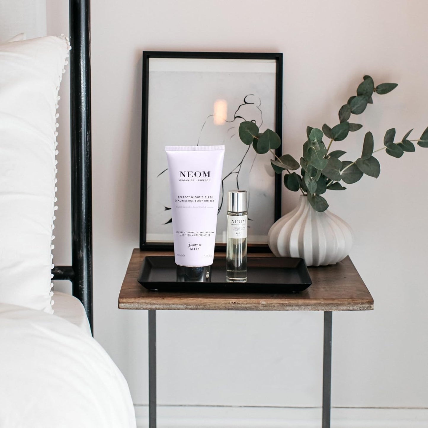 Neom skincare products on a bedside table with a framed picture and plant in the background.