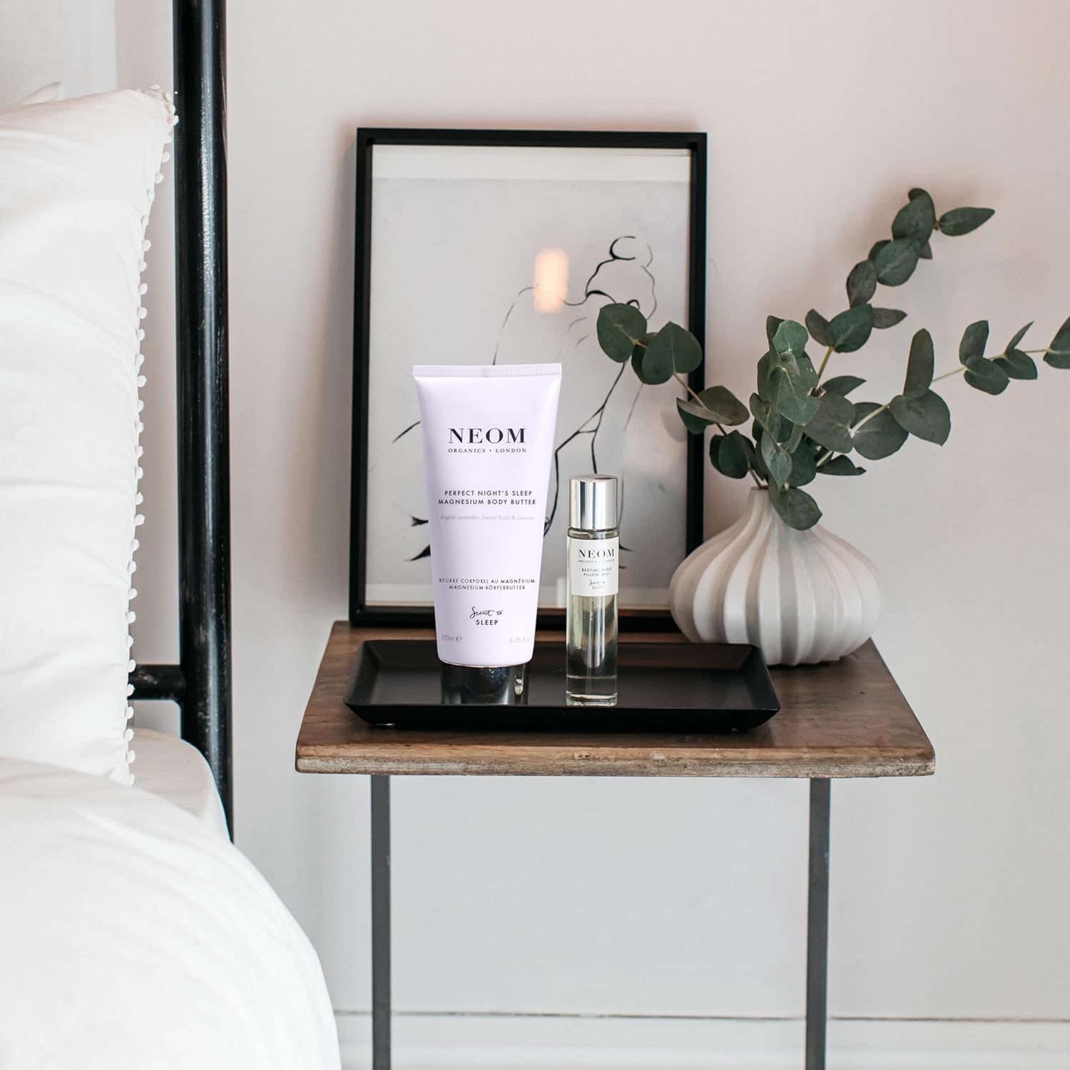 Neom skincare products on a bedside table with a framed picture and plant in the background.