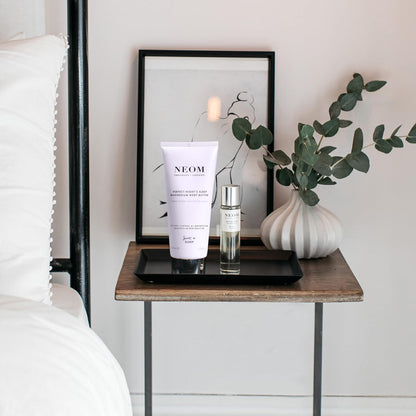 Neom skincare products on a bedside table with a framed picture and plant in the background.