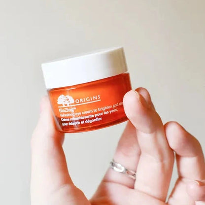 Hand holding a small jar of Origins eye cream against a plain background