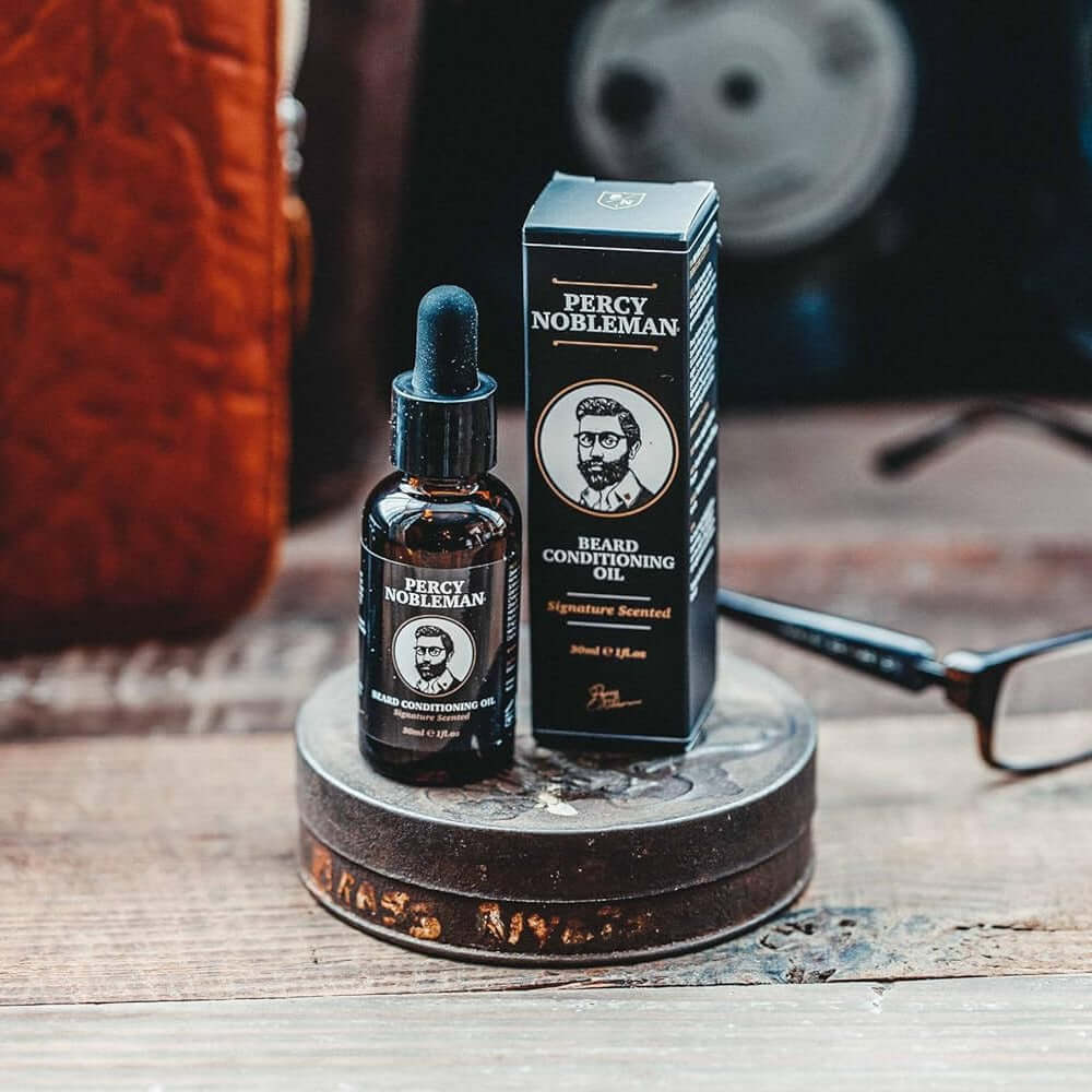 Percy Nobleman beard conditioning balm bottle and packaging on a wooden surface with glasses and a camera in the background.