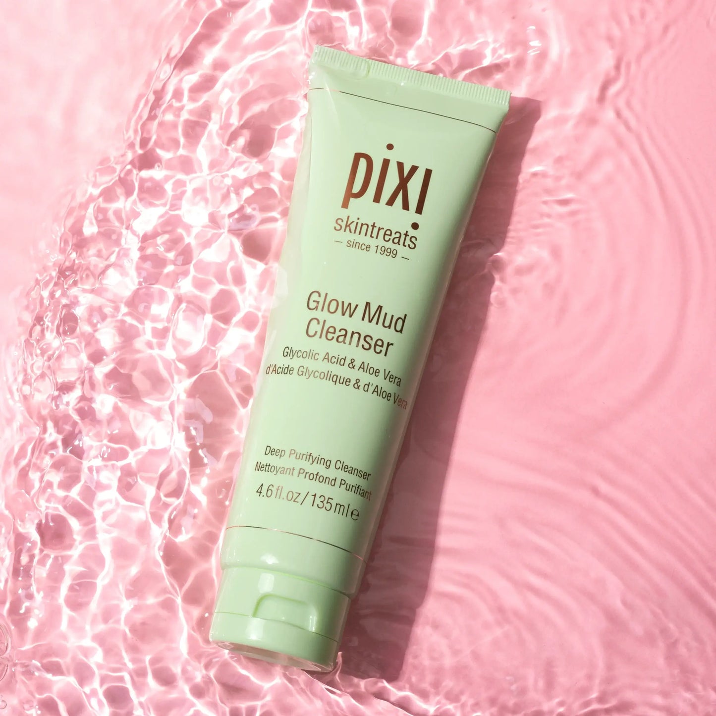 PIXI Glow Mud Cleanser 135ml tube on pink surface with water, enriched with Glycolic Acid and Aloe Vera for radiant skin.