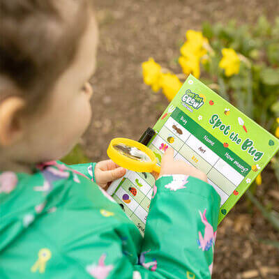Child using a magnifying glass to explore the PlayWorks Spot The Bugs Checklist in a garden with flowers.