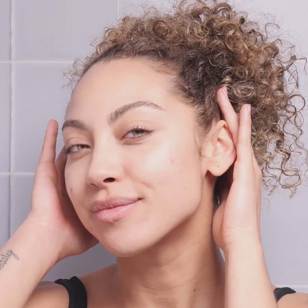 Model showcasing clear skin while using The INKEY List Fulvic Acid Cleanser in a bright bathroom setting.