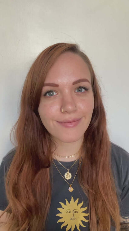 Woman with long red hair, wearing a sun graphic t-shirt and layered jewelry, smiling at the camera.