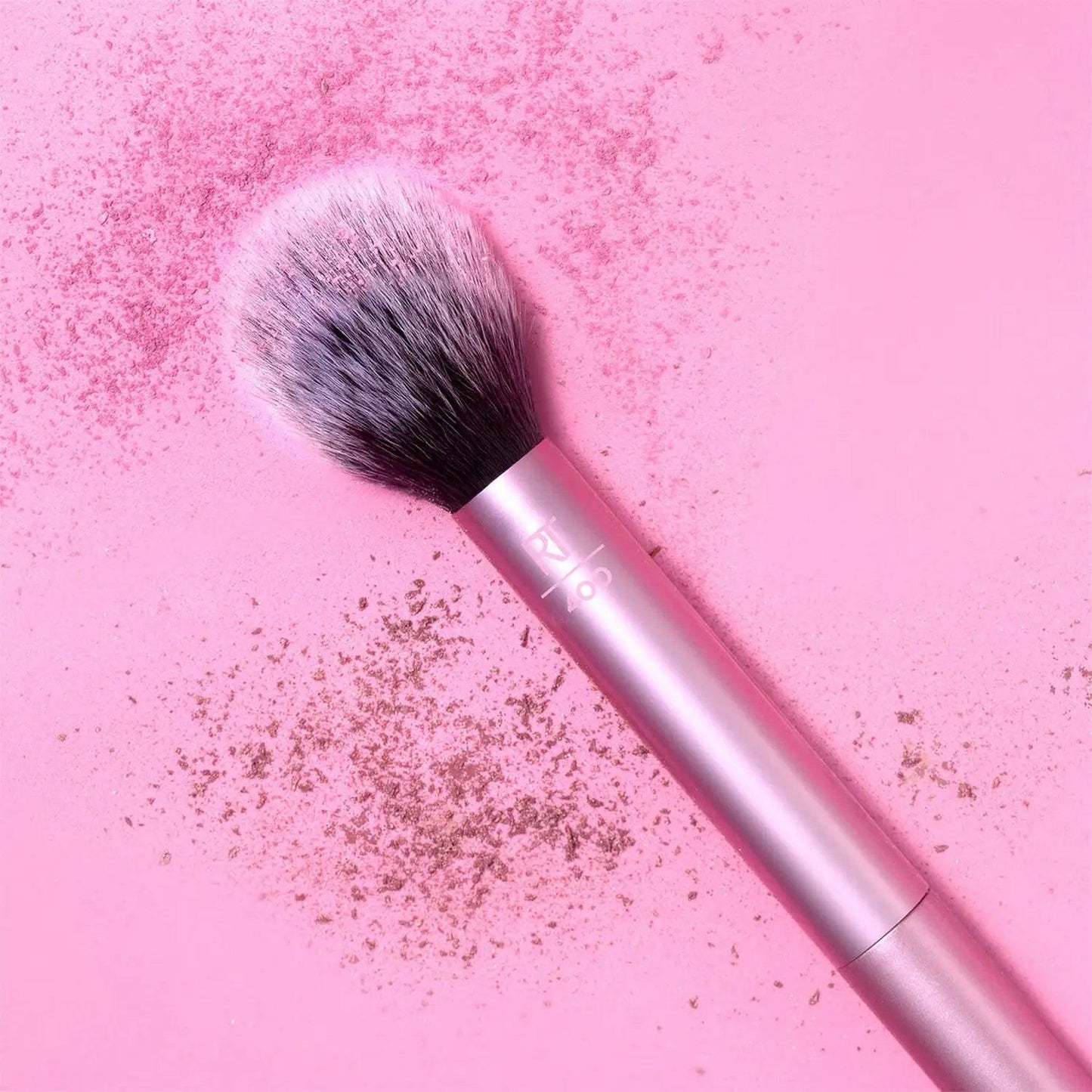 Real Techniques Blush Brush on a pink background with blush powder, showcasing its fluffy head for flawless application.