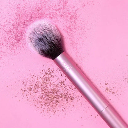 Real Techniques Blush Brush on a pink background with blush powder, showcasing its fluffy head for flawless application.