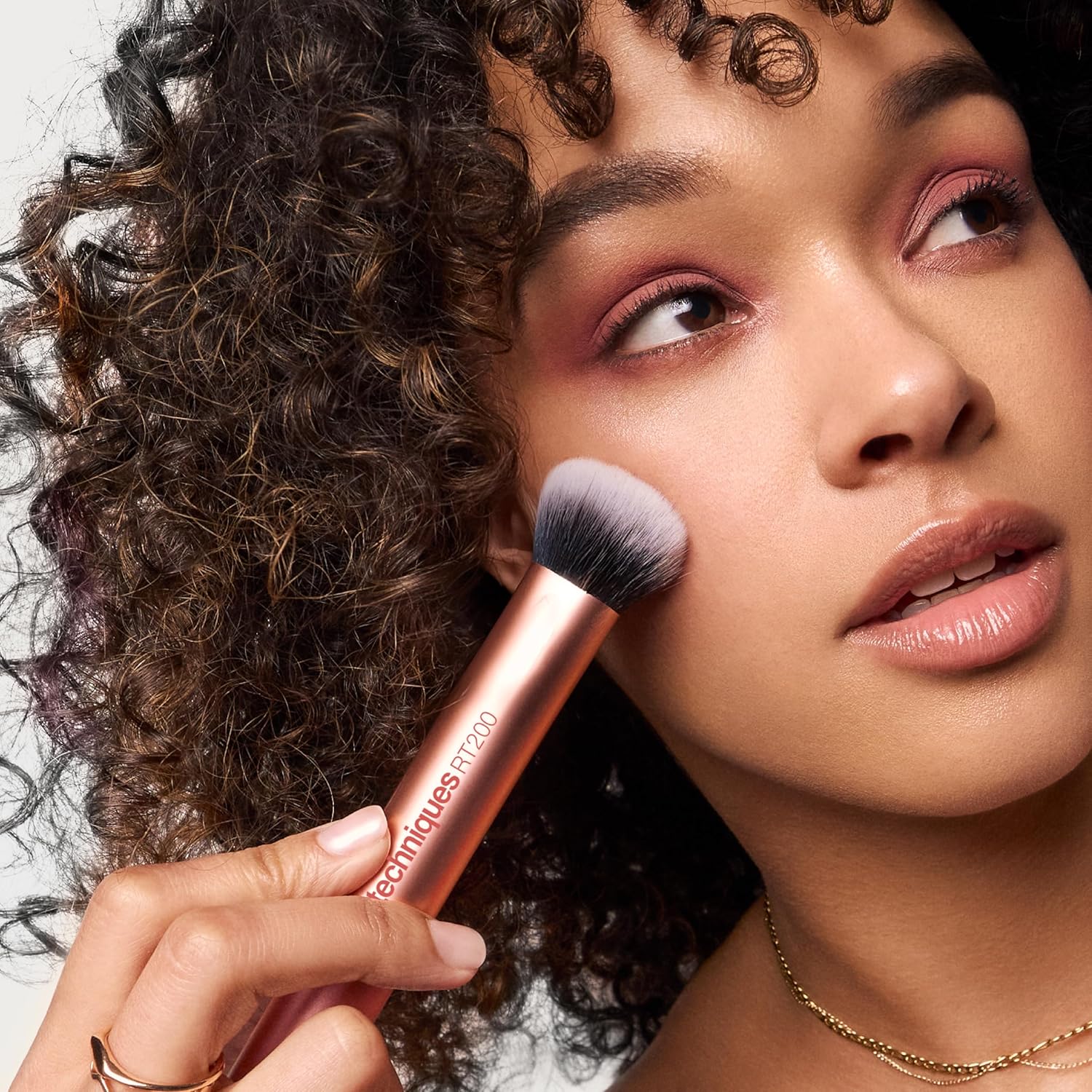 Real Techniques Woman applying makeup with a brush, wearing a gold necklace.