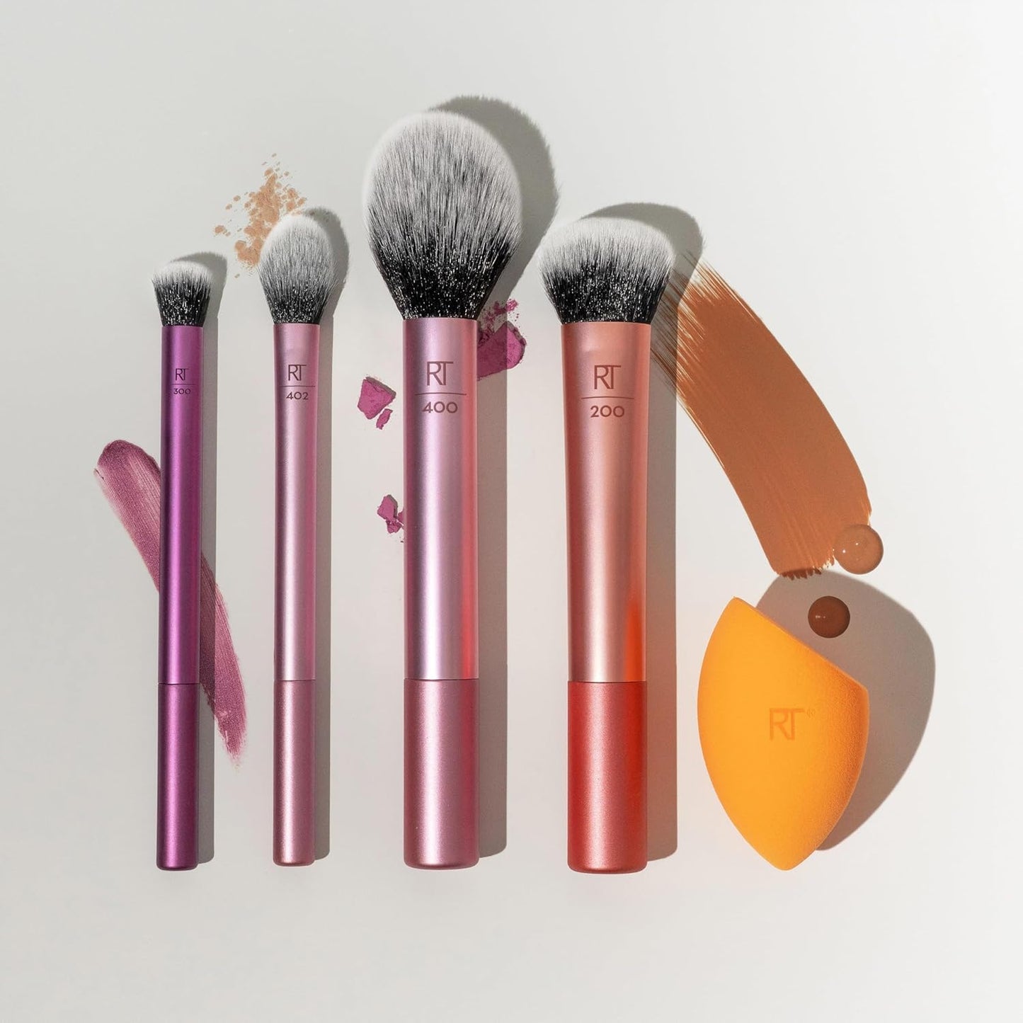Real Techniques Makeup brushes with pink and orange handles and an orange sponge applicator on a light background.