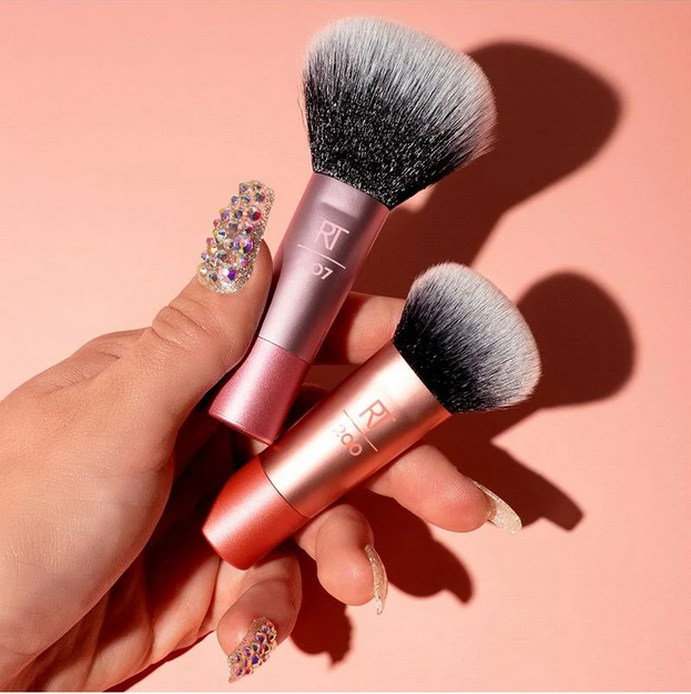 Two makeup brushes held by a hand with decorative nails on a pink background