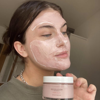 Person applying pink clay mask to face with Revolution Skincare jar in foreground