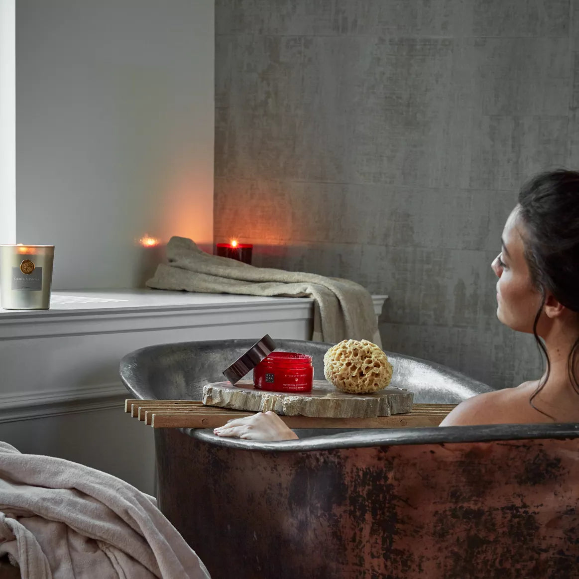 Woman in a bathtub with a candle, sponge, and container on a wooden board.