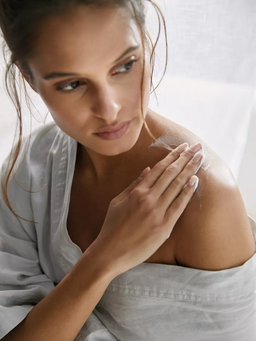 Woman applying a skincare product to her shoulder with a neutral background