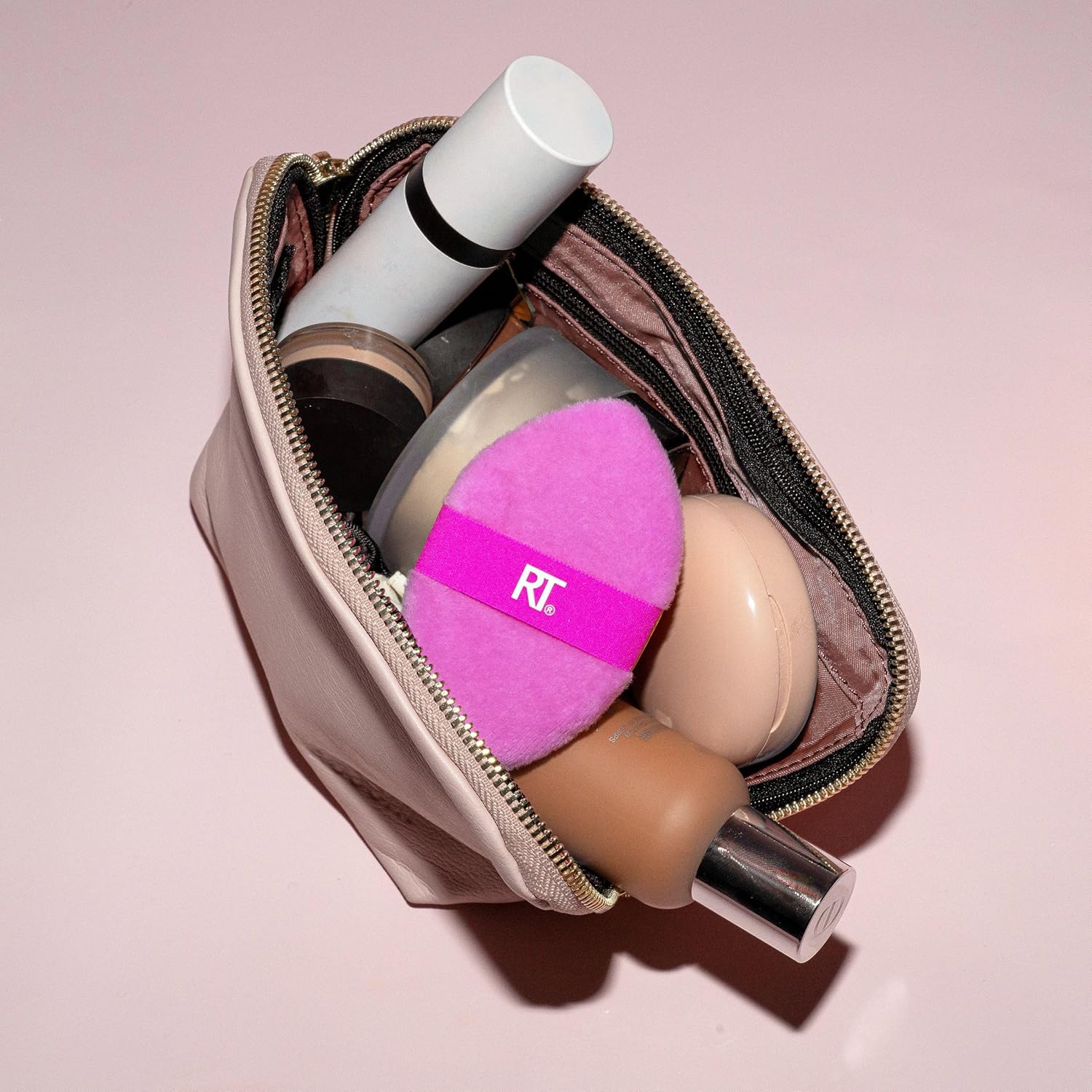 Makeup products including a pink puff with 'RT' branding in an open makeup bag on a pink background.