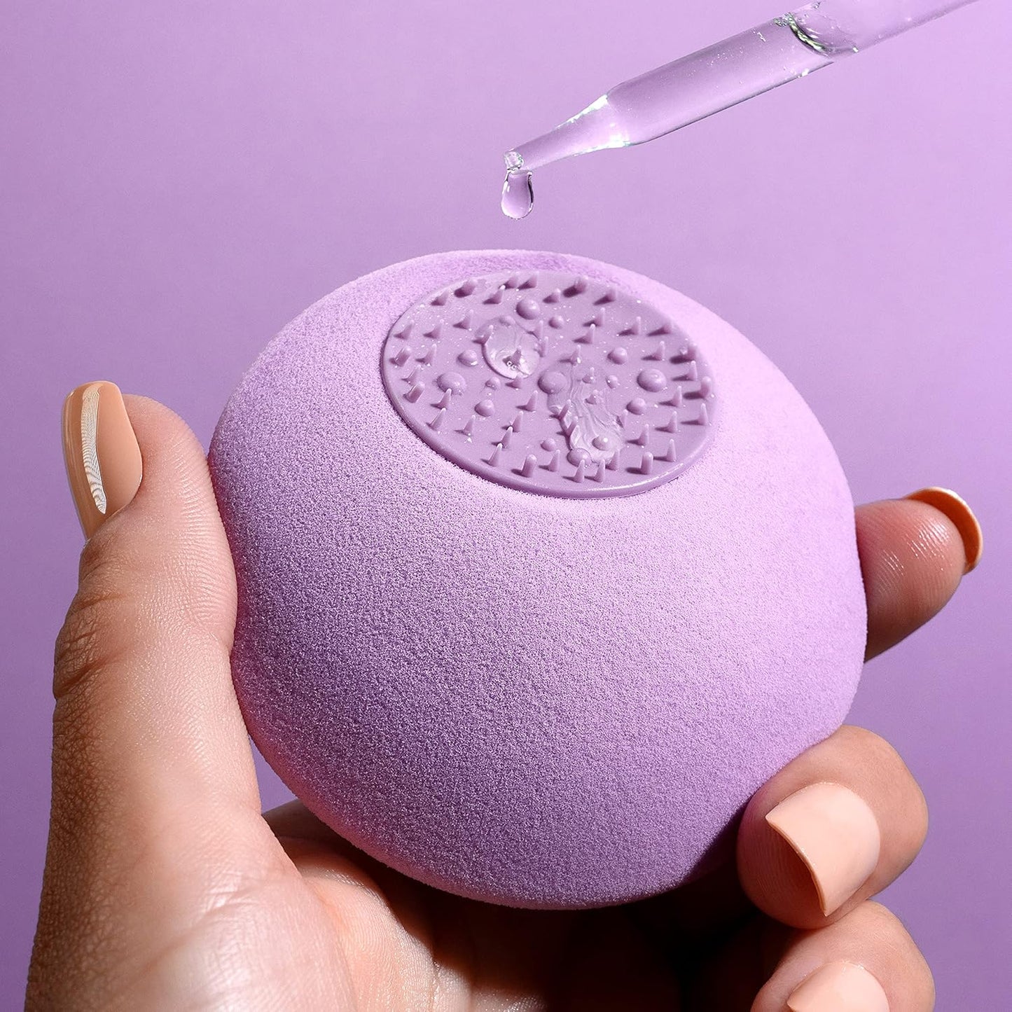 Purple textured ball held by a hand with a dropper of liquid above it on a purple background