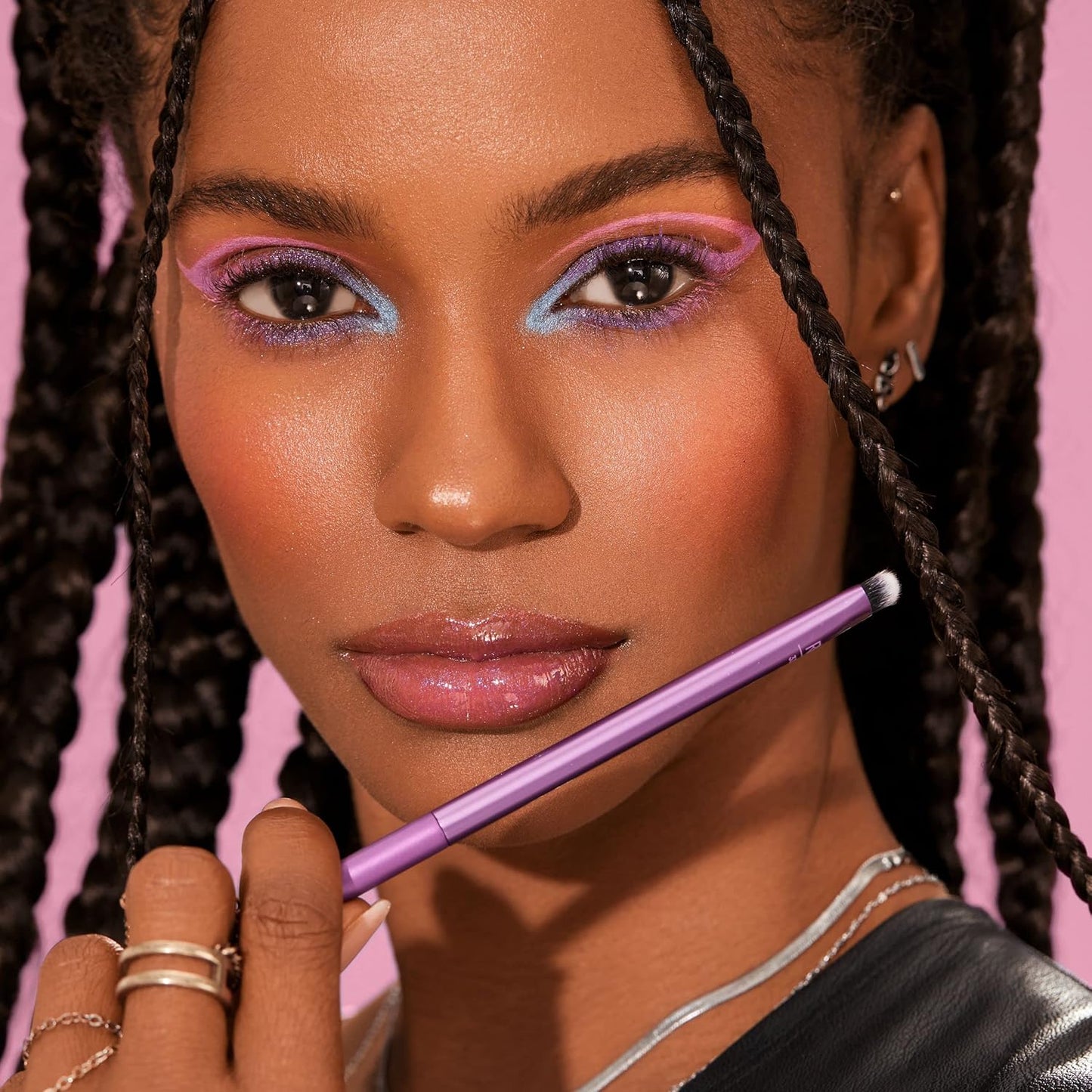 Woman with colorful makeup holding a purple makeup pencil against a pink background