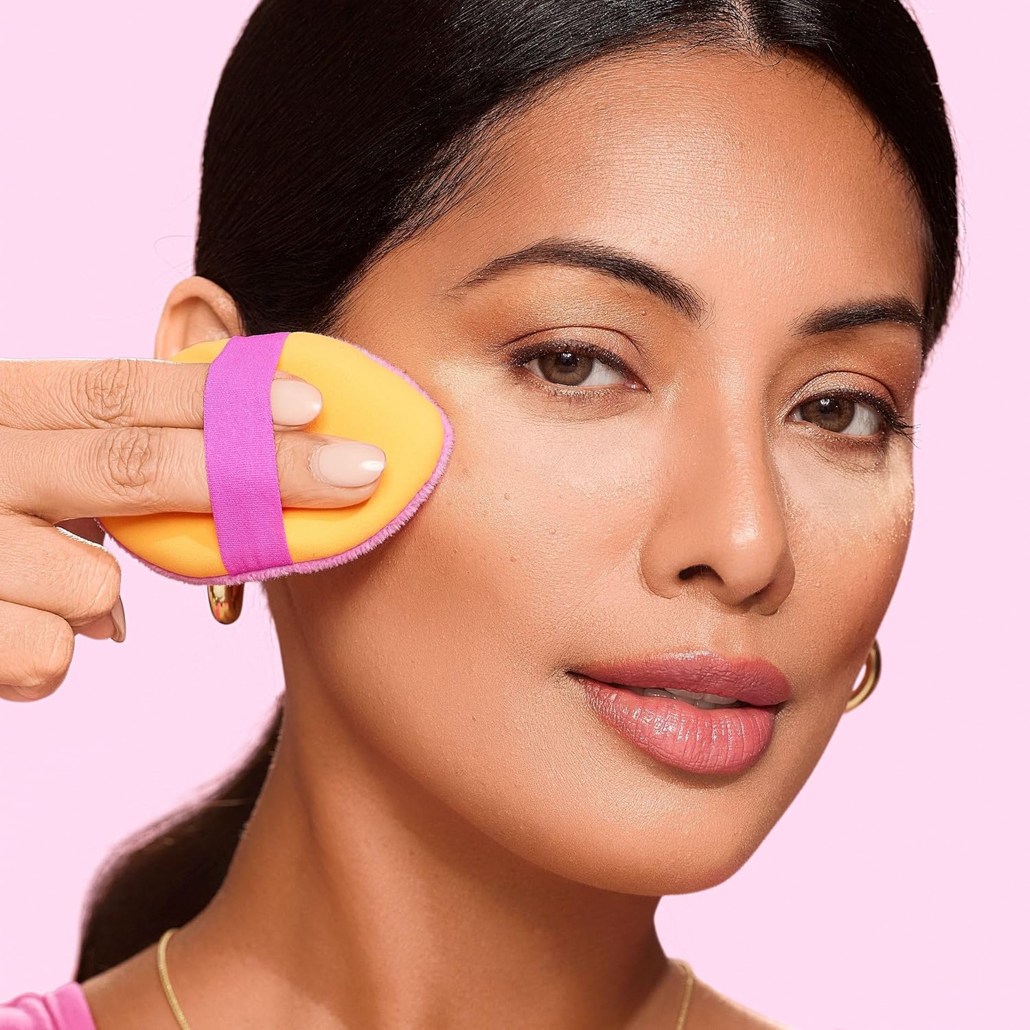 Woman using a beauty blender on her face against a pink background