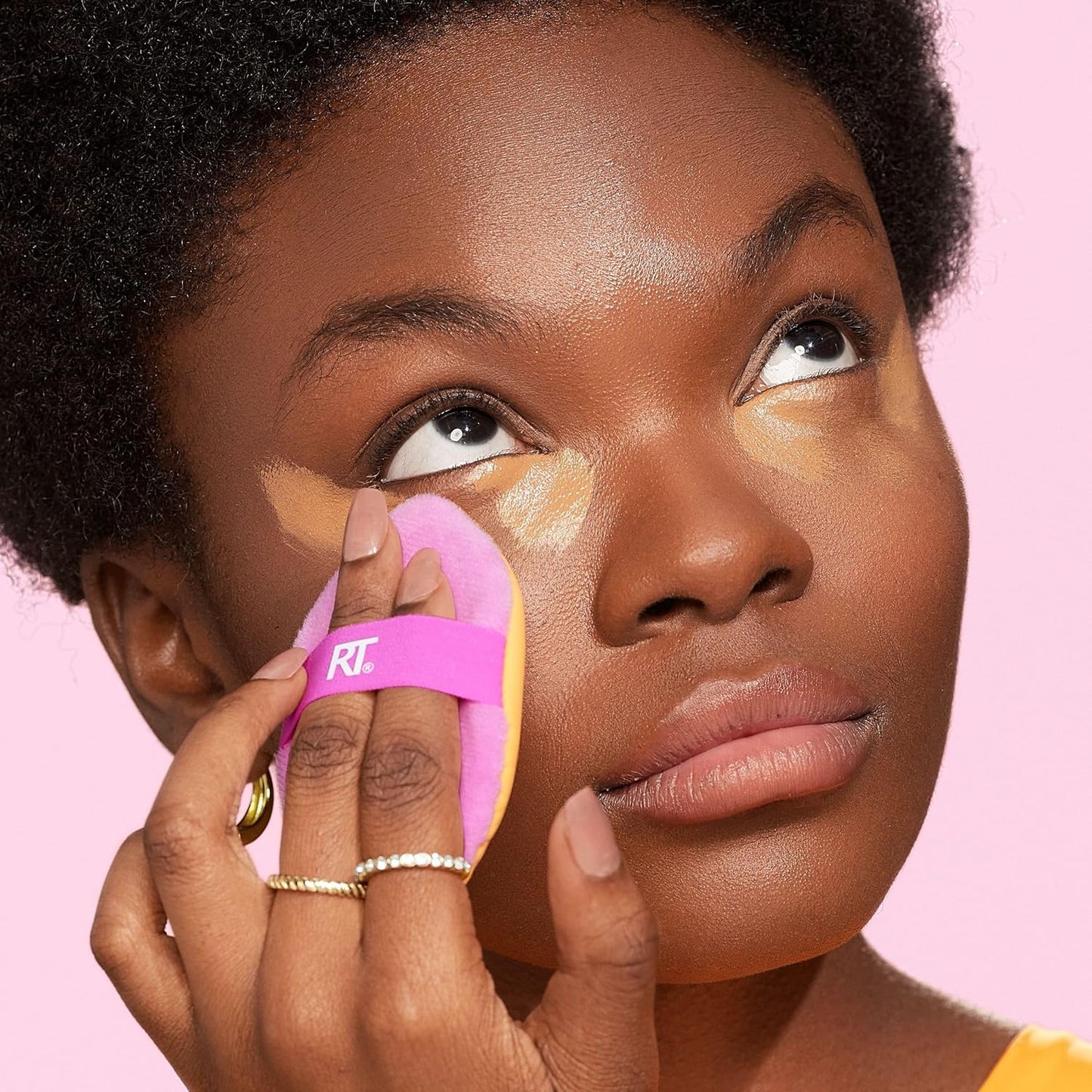 Woman applying makeup with a pink sponge applicator on a pink background