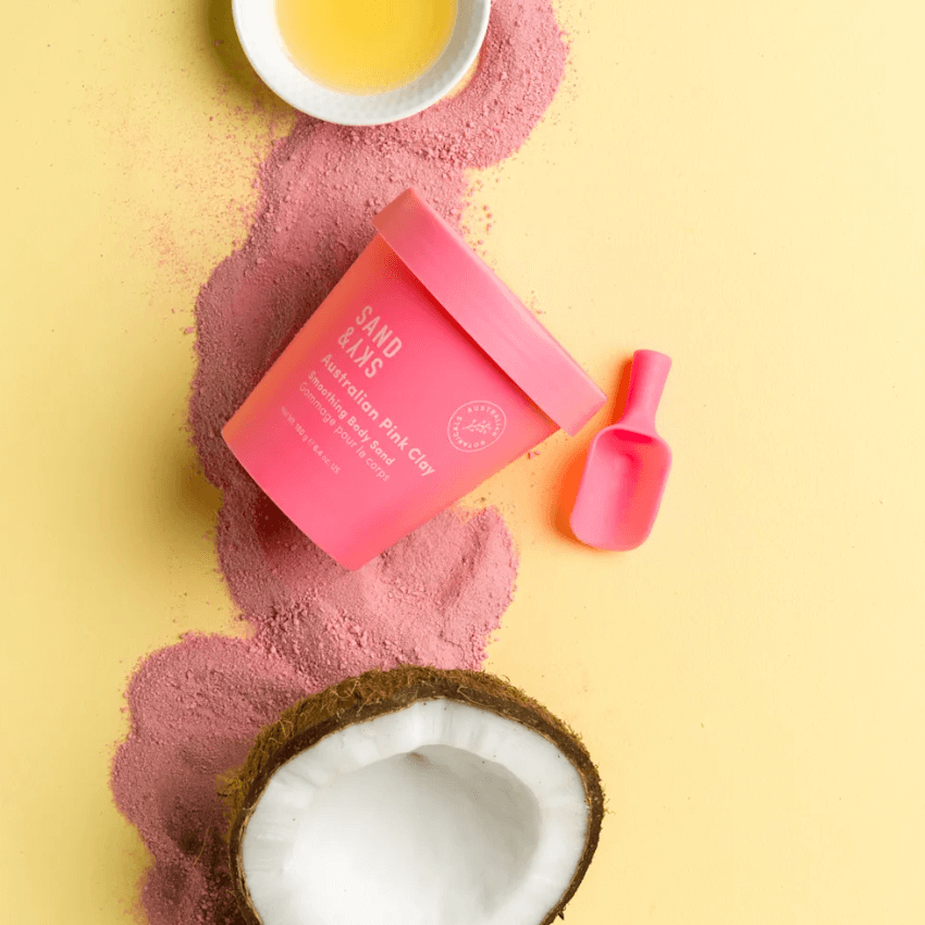 Sand & Sky Australian Pink Clay Smoothing Body Sand on a yellow background with coconut and essential oil bowl.