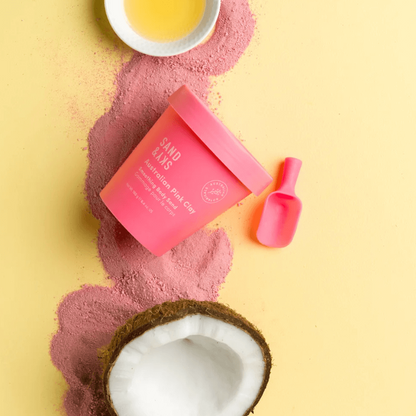 Sand & Sky Australian Pink Clay Smoothing Body Sand on a yellow background with coconut and essential oil bowl.