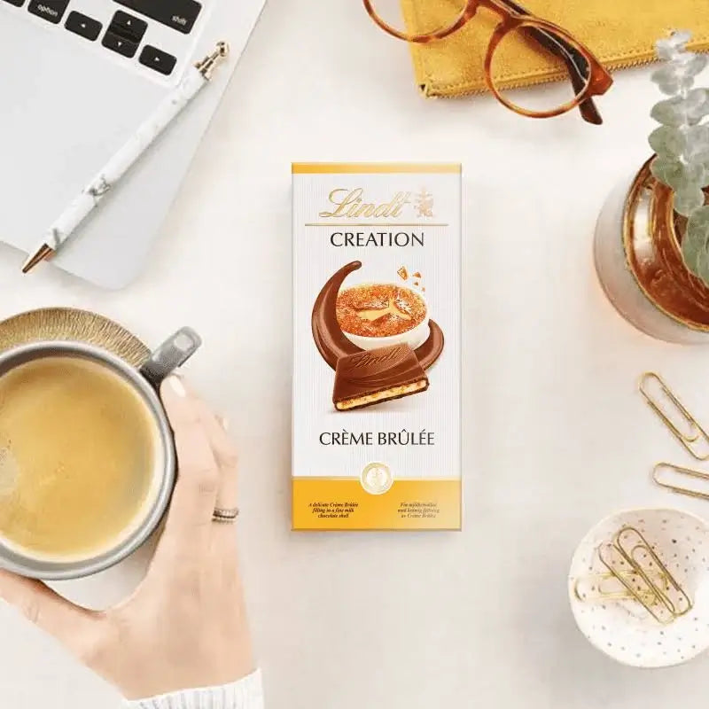 Lindt Crème Brélé chocolate bar with a cup of coffee and office items on a desk.