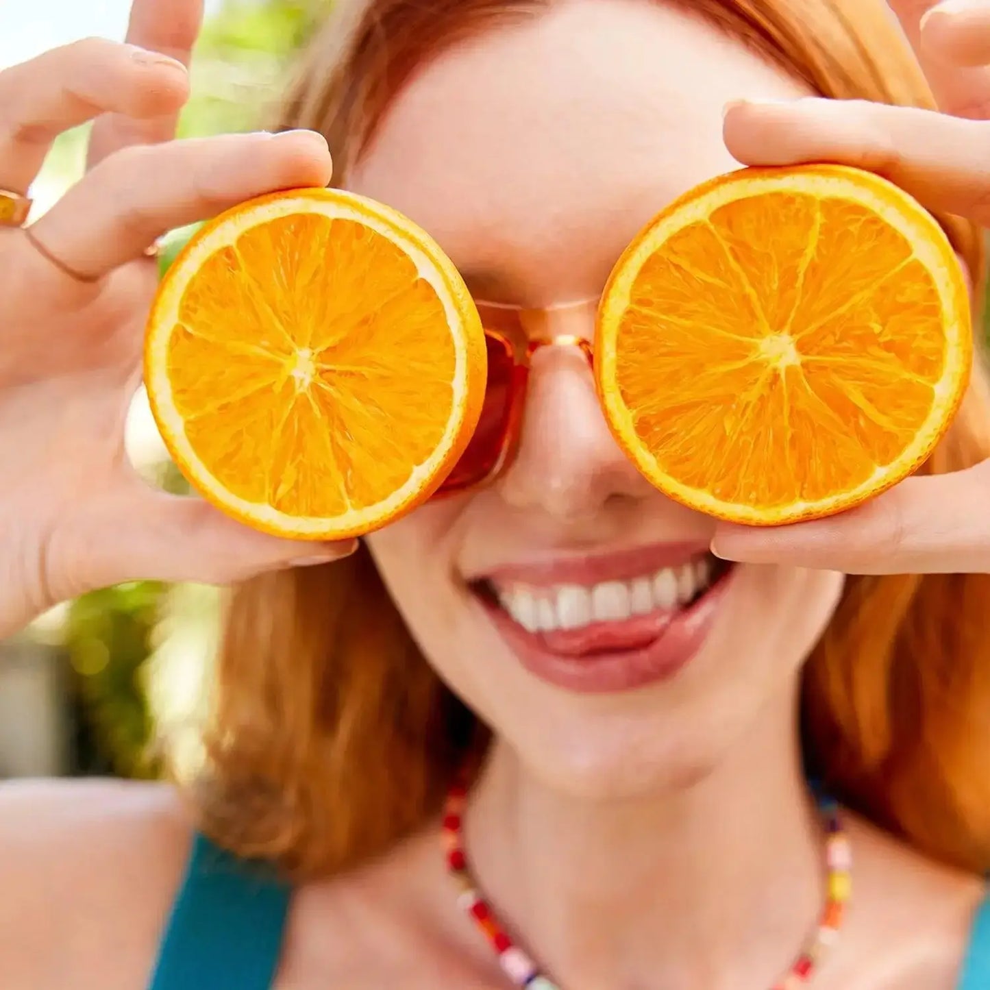 First Aid Beauty Smiling woman holding orange slices in front of her eyes, wearing colorful sunglasses and a beaded necklace.