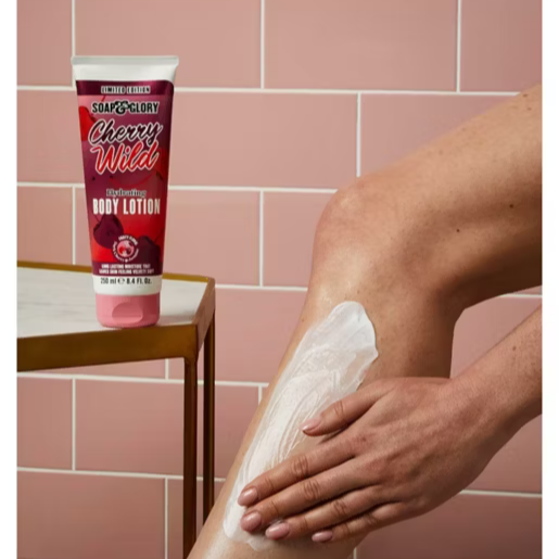 Person applying body lotion to their leg with a Cherry Wild tube of lotion on a pink tiled wall background.