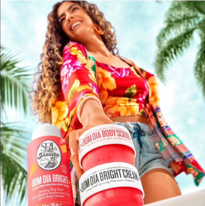Sol de Janeiro Bom Dia Bright Clarifying AHA BHA Body Wash with body scrub and cream by a smiling woman in floral attire.