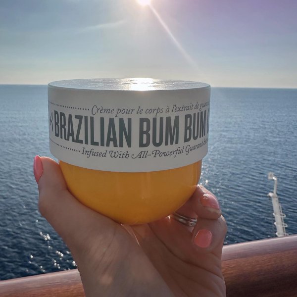 Hand holding a jar of Sol de Janeiro 'Brazilian Bum Bum' cream with a body of water in the background