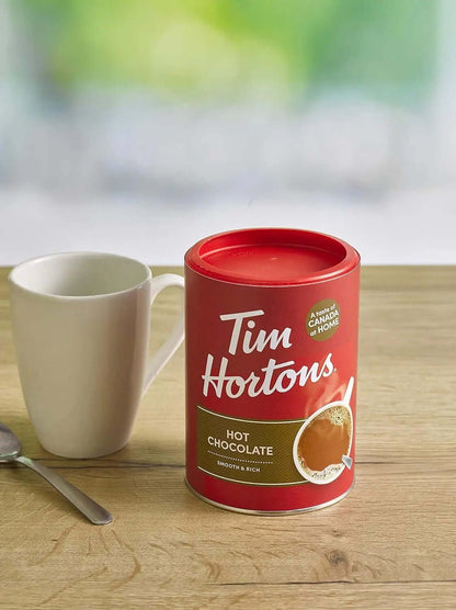 Tim Hortons Hot Chocolate Mix 500g can beside a cup on a wooden table, perfect for classic hot chocolate lovers.