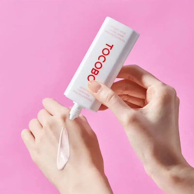 TOCOBO Vita Tone Up Sun Cream SPF50+ being applied to hand, showcasing smooth texture, with pink background.
