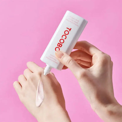 TOCOBO Vita Tone Up Sun Cream SPF50+ being applied to hand, showcasing smooth texture, with pink background.
